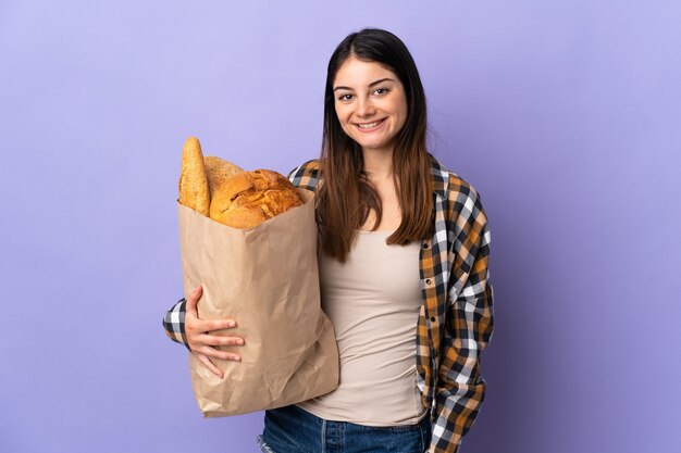 Jovem segurando uma sacola cheia de pães isolados no roxo e sorrindo muito