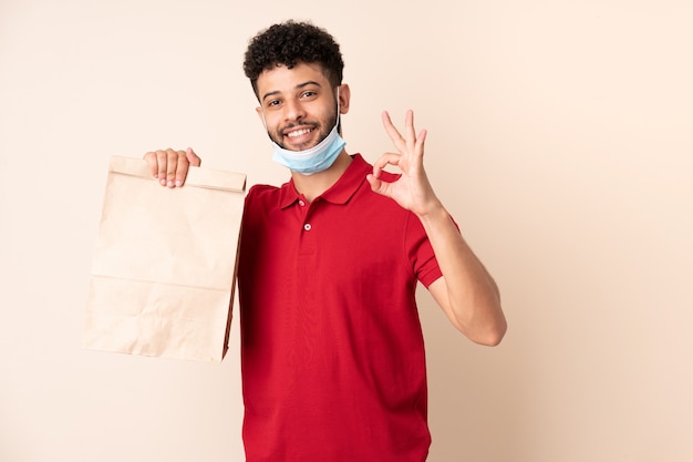 Jovem segurando uma sacola de comida para viagem ___ mostrando sinal de ok com os dedos