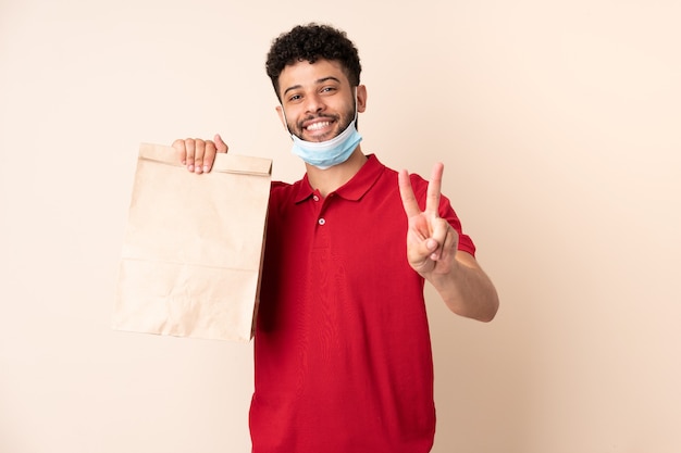 Jovem segurando uma sacola de comida para viagem ___ sorrindo e mostrando sinal de vitória