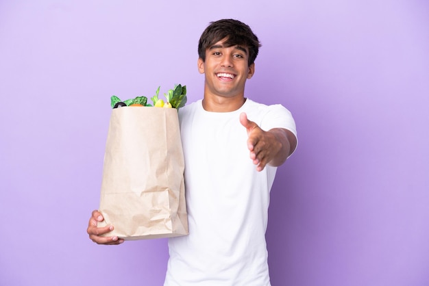 Jovem segurando uma sacola de compras isolada em fundo roxo, apertando as mãos para fechar um bom negócio