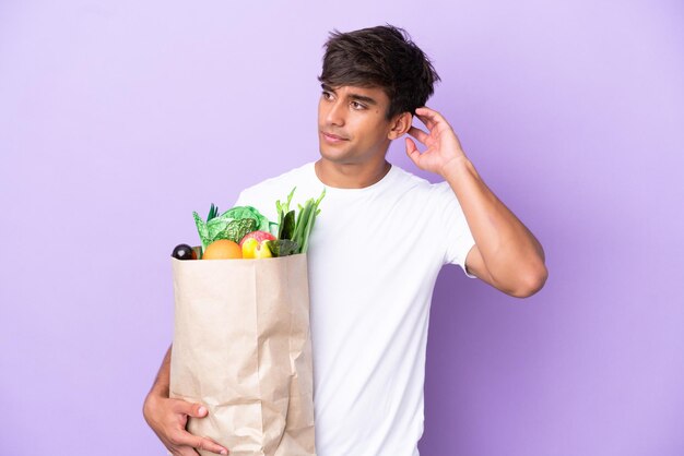 Jovem segurando uma sacola de compras isolada em fundo roxo com dúvidas