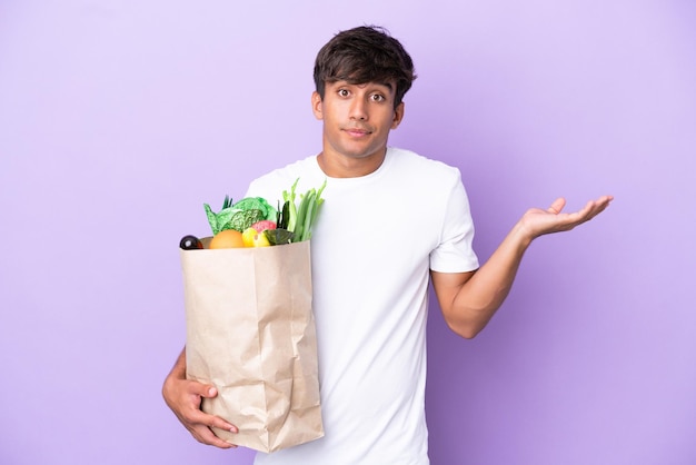 Jovem segurando uma sacola de compras isolada no fundo roxo com dúvidas ao levantar as mãos