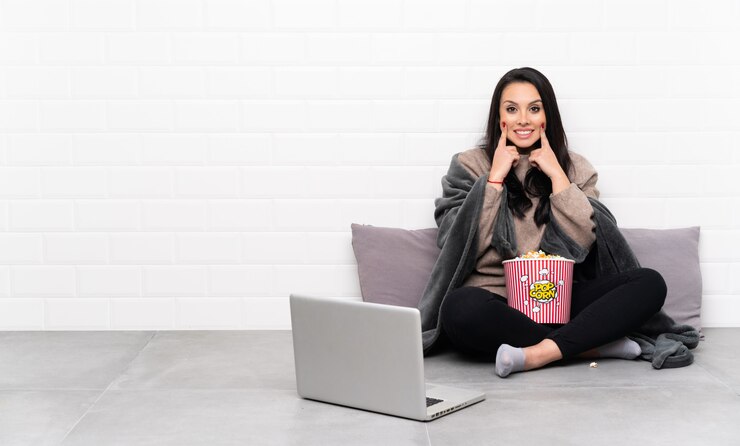 Foto jovem, segurando uma tigela de pipocas e mostrando um filme em um laptop, sorrindo com uma expressão feliz e satisfeito