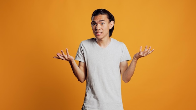 Jovem sem noção fazendo sinal de não sei no estúdio, agindo incerto e inseguro sobre a resposta à pergunta. Um cara asiático sendo despreocupado e casual, agindo de forma duvidosa sobre o pano de fundo laranja.