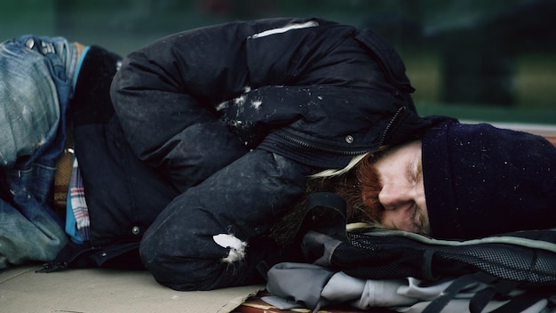 Jovem sem-teto bêbado tentando dormir em papelão no banco na calçada