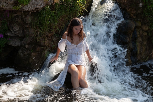 Jovem seminua em um vestido branco aprecia o frescor e o frescor dos jatos d'água da cachoeira