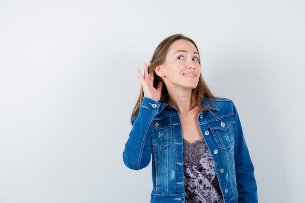 Jovem senhora com a mão atrás da orelha na blusa, jaqueta jeans e bonito. vista frontal.