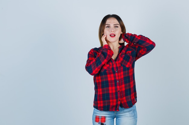 Jovem senhora com a mão na bochecha, tocando seu pescoço em uma camisa xadrez e olhando perplexa, vista frontal.