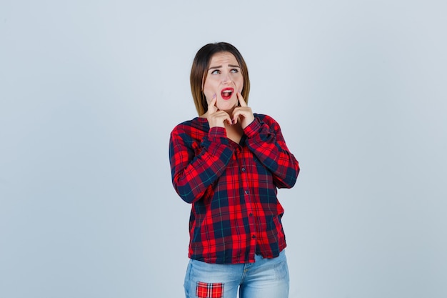 Jovem senhora com as mãos nas bochechas, abrindo a boca em uma camisa xadrez, jeans e parecendo descontente, vista frontal.