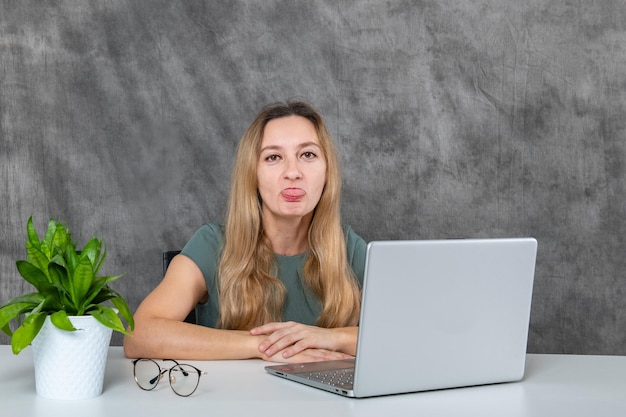 Jovem senhora com cabelo loiro em vestido cinzento poses impressionantes perto de laptop e flor verde