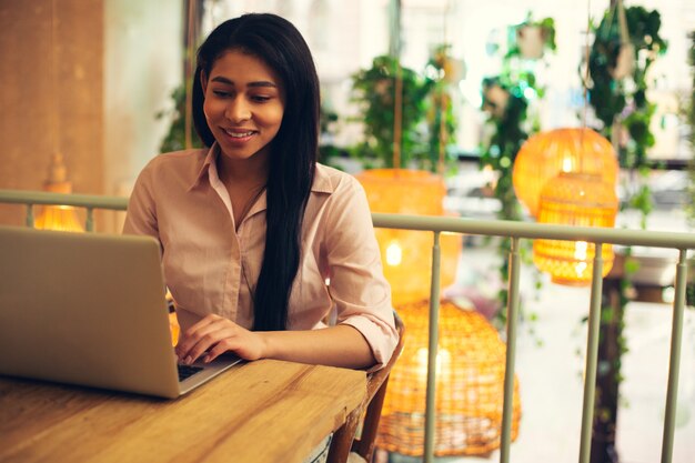 Jovem senhora de cabelos escuros sentada à mesa em um café confortável e sorrindo enquanto olha para a tela de um laptop moderno