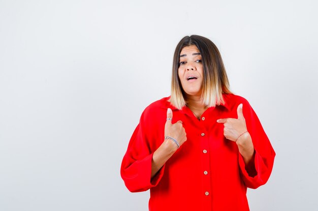 Jovem senhora de camisa vermelha grande, apontando para si mesma fazendo uma pergunta e parecendo animada, vista frontal.