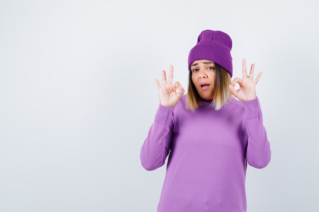 Jovem senhora de suéter roxo, gorro mostrando o gesto de ok e olhando perplexa, vista frontal.