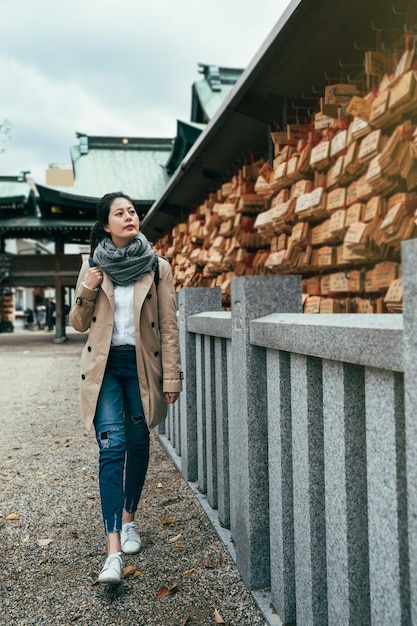 jovem senhora de viagens asiáticas andando no templo japonês em osaka através da parede de oração. placa de madeira cheia de desejos e esperanças usando corda vermelha pendurada. menina olhando para a fortuna curiosamente.