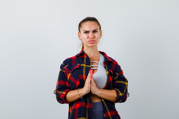 Jovem senhora em top, camisa xadrez mostrando gesto de meditação e olhando esperançosa, vista frontal.