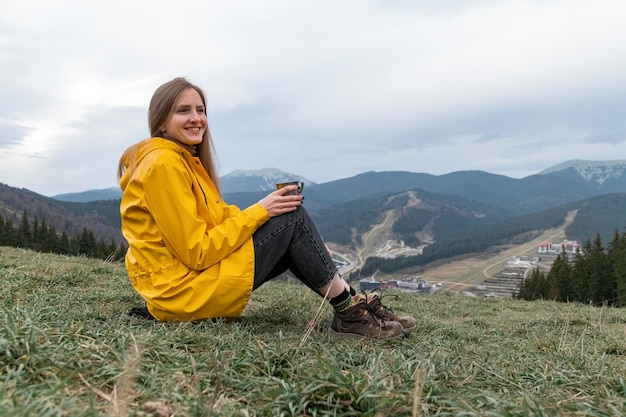 Jovem senta-se na encosta bebendo chá e sorrindo Retrato de turista em jaqueta amarela nas montanhas