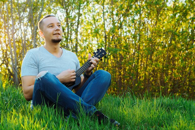 Jovem senta-se tocando ukulele no parque.