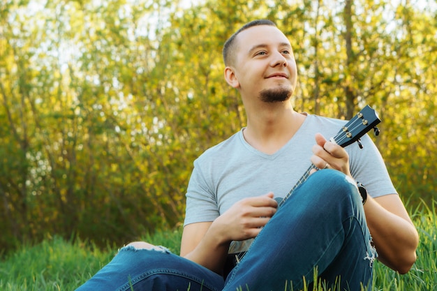 Jovem senta-se tocando ukulele no parque.