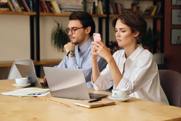 Jovem sentada à mesa com laptop e xícara de café pensativamente usando celular colegas de trabalho trabalhando no escritório moderno