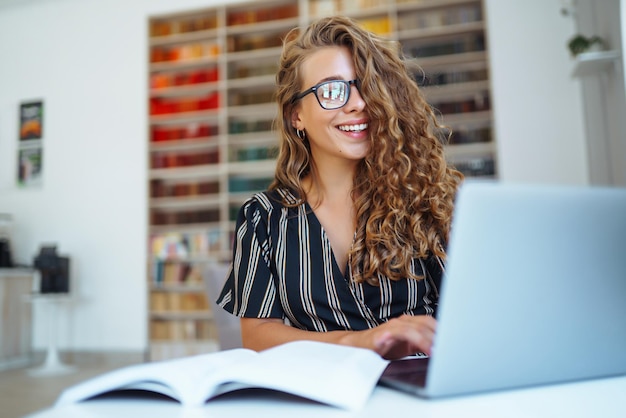 Jovem sentada com um laptop de livros na biblioteca e tomando notas de aprendizagem Conceito de educação