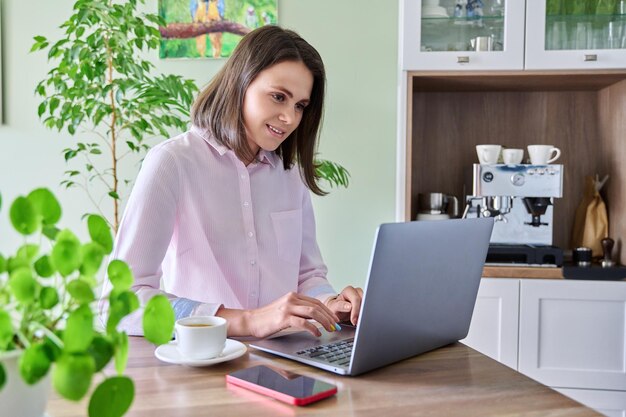 Jovem sentada em casa na cozinha usando laptop bebendo café da manhã Estilo de vida freelancer tecnologia e trabalho lazer conceito de pessoas de 2530 anos