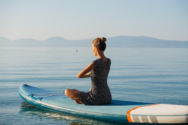 Jovem sentada em posição de lótus meditando na prancha de sup flutuando na água calma do mar matinal