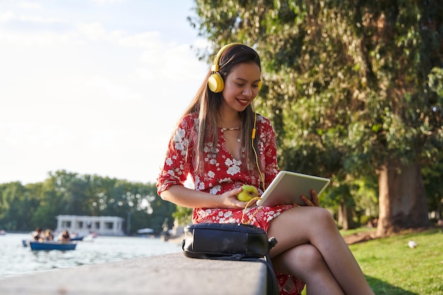 Jovem sentada em um parque comendo uma maçã enquanto olha para seu tablet com seus fones de ouvido