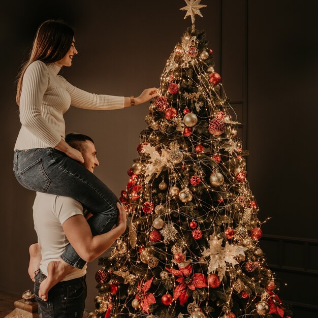 Jovem sentada nas costas de um homem e decorando uma árvore de natal pendura enfeites