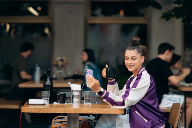 Jovem sentada no café ao ar livre e tomando café segurando o smartphone