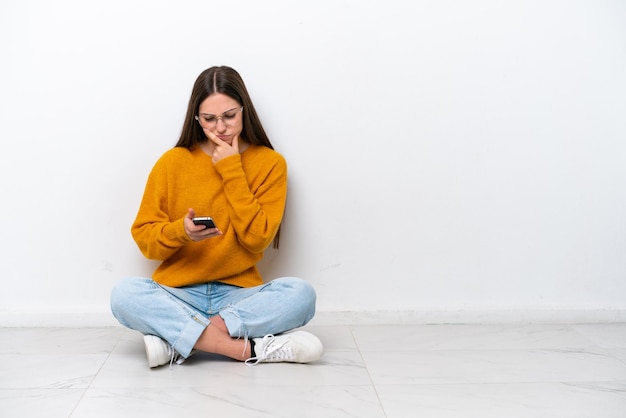 Jovem sentada no chão isolada no fundo branco pensando e enviando uma mensagem