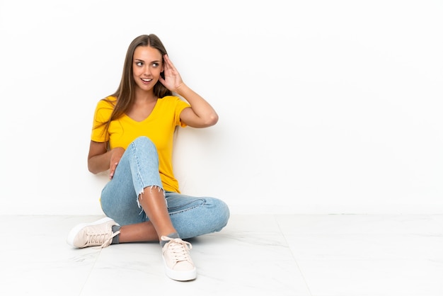 Jovem sentada no chão ouvindo algo colocando a mão na orelha