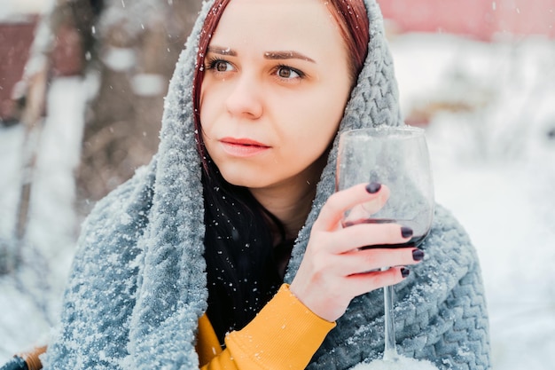Jovem sentada no quintal e bebendo vinho tinto em clima de neve Fêmea embrulhada em xadrez cinza sentado na rua com álcool na temporada de inverno