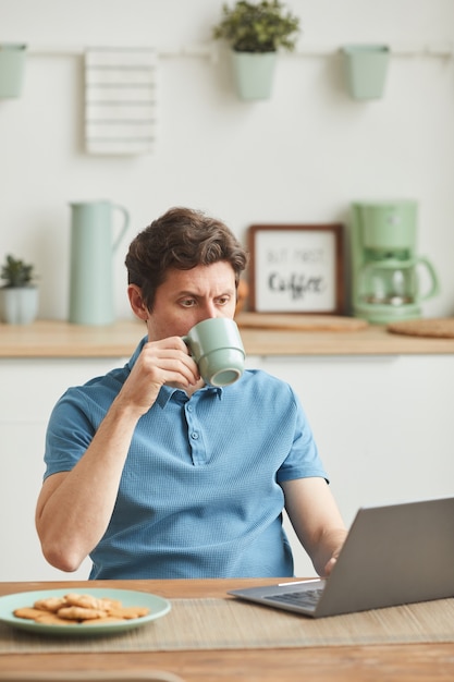 Jovem sentado à mesa olhando para o monitor do laptop e bebendo café na cozinha doméstica