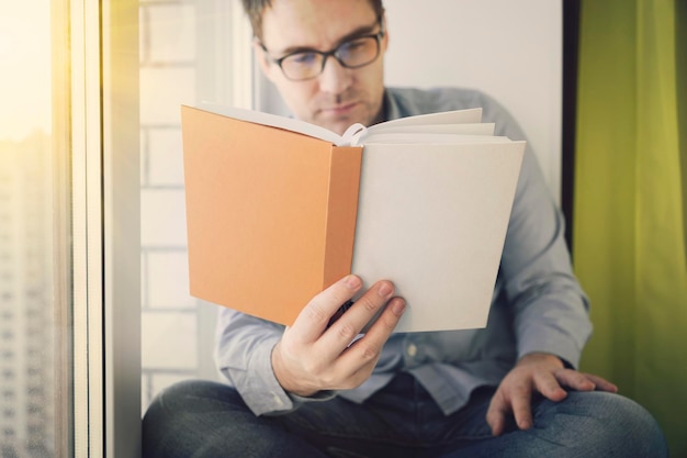 Jovem sentado ao lado de uma janela lendo um livro e aproveitando seu tempo de lazer