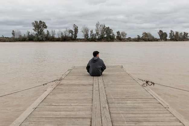 Foto jovem sentado em um píer próximo a um rio, olhando e descansando em um céu nublado