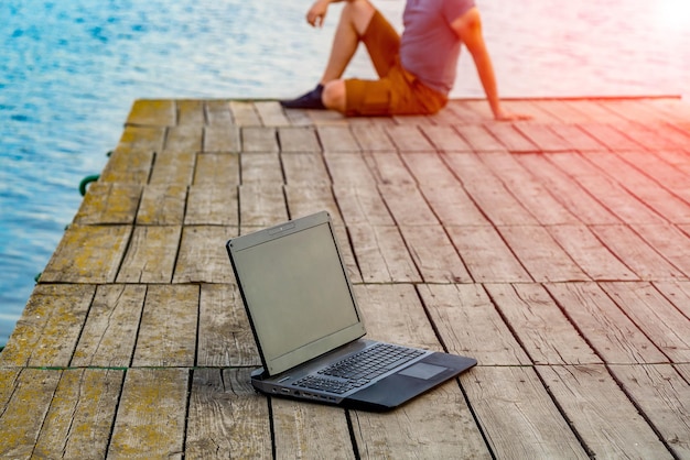 Jovem sentado em um píer usando seu laptop Trabalho e férias