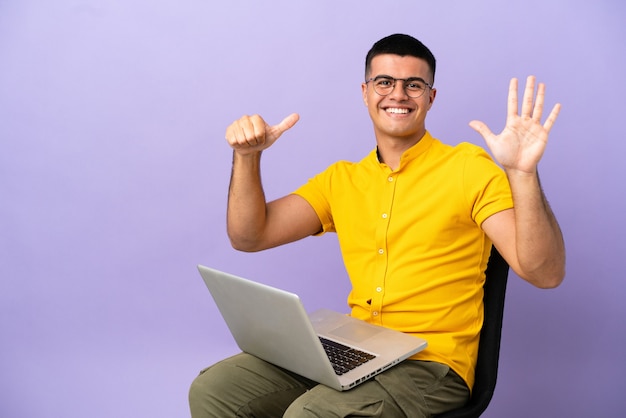 Jovem sentado em uma cadeira com laptop, contando seis com os dedos