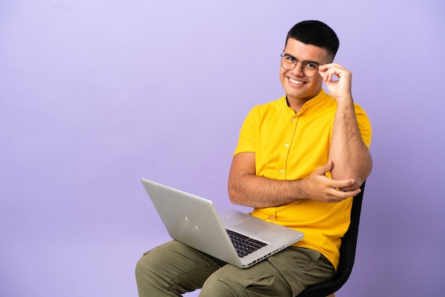 Jovem sentado em uma cadeira com laptop de óculos e feliz