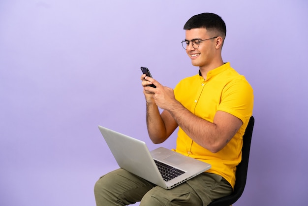 Jovem sentado em uma cadeira com laptop enviando uma mensagem com o celular