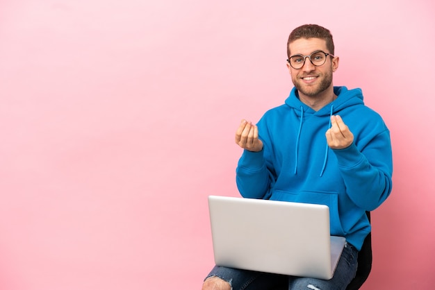 Jovem sentado em uma cadeira com laptop fazendo gesto de dinheiro