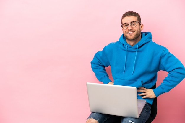 Jovem sentado em uma cadeira com laptop posando com os braços na cintura e sorrindo