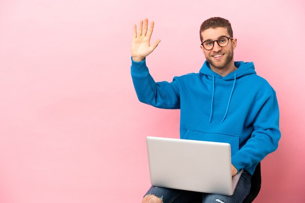 Jovem sentado em uma cadeira com laptop saudando com a mão com expressão feliz