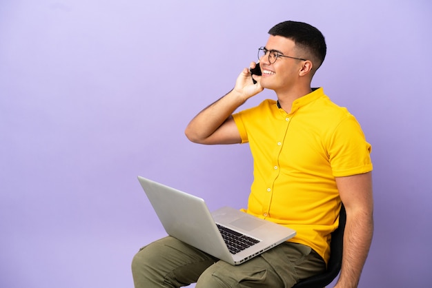 Jovem sentado em uma cadeira com um laptop, conversando com alguém ao telefone celular