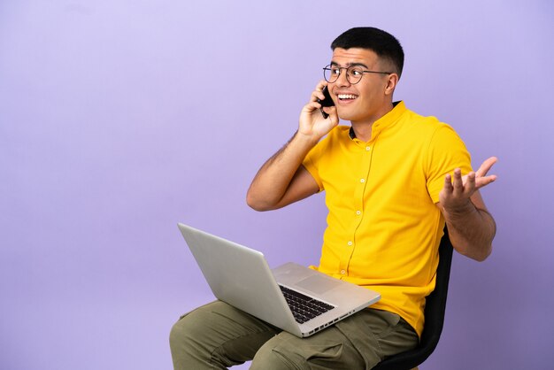 Jovem sentado em uma cadeira com um laptop, conversando com alguém ao telefone celular