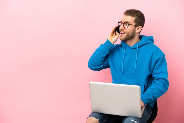 Jovem sentado em uma cadeira com um laptop, conversando com o celular