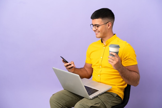 Jovem sentado em uma cadeira com um laptop segurando um café para levar e um celular