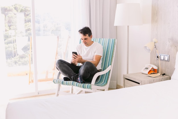 Jovem sentado em uma cadeira de balanço em seu quarto com as pernas cruzadas olhando para o celular com luz entrando pela janela Conceito de conexão de férias de trabalho e smartphone