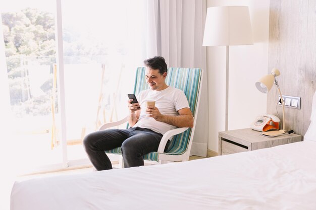 Jovem sentado em uma cadeira de balanço em seu quarto olhando para o celular e segurando uma bebida com a luz entrando pela janela Conceito de trabalhar férias café café da manhã