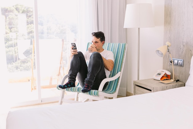 Jovem sentado em uma cadeira de balanço em seu quarto olhando para o celular e tomando um café com a luz entrando pela janela Conceito de conexão de férias de trabalho e smartphone