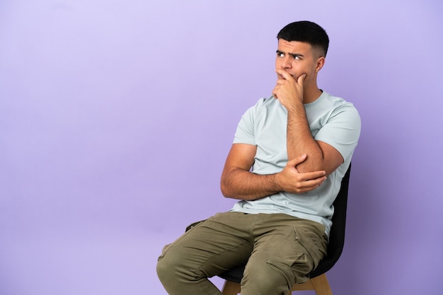 Jovem sentado em uma cadeira sobre um fundo isolado, tendo dúvidas e com expressão facial confusa